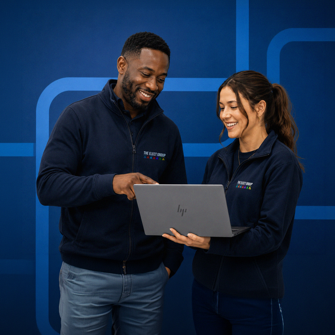 Two people wearing matching navy jackets with 'The Elect Group' logo looking at a laptop and smiling, standing in front of a blue background with geometric design.