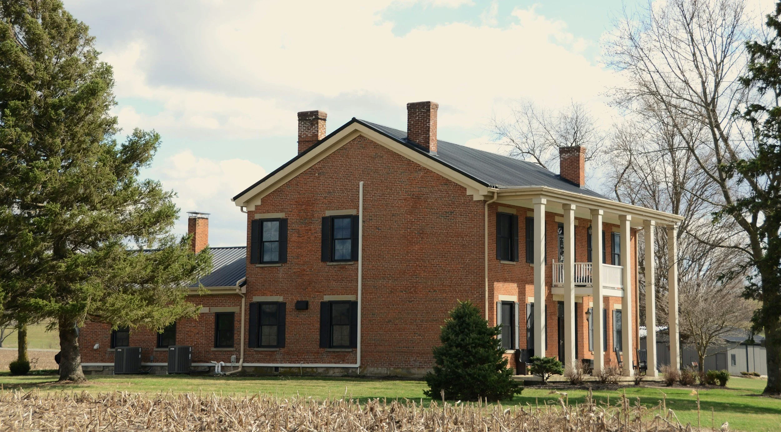 A large brick house with tall white columns on a porch, black window shutters, multiple chimneys, and surrounded by trees and a grassy yard.