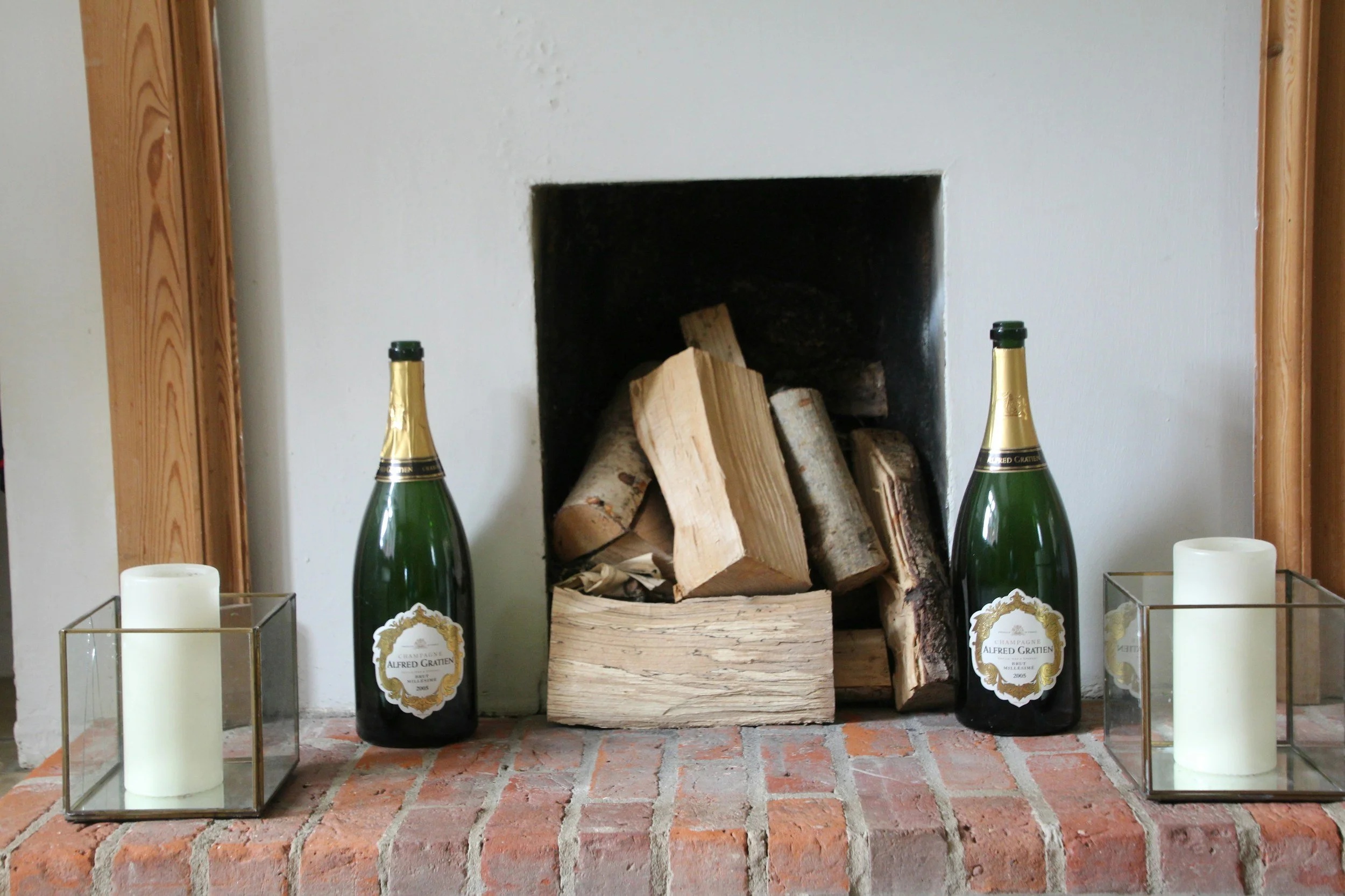 Fireplace with chopped wood, two bottles of champagne, and two white candles in glass holders.