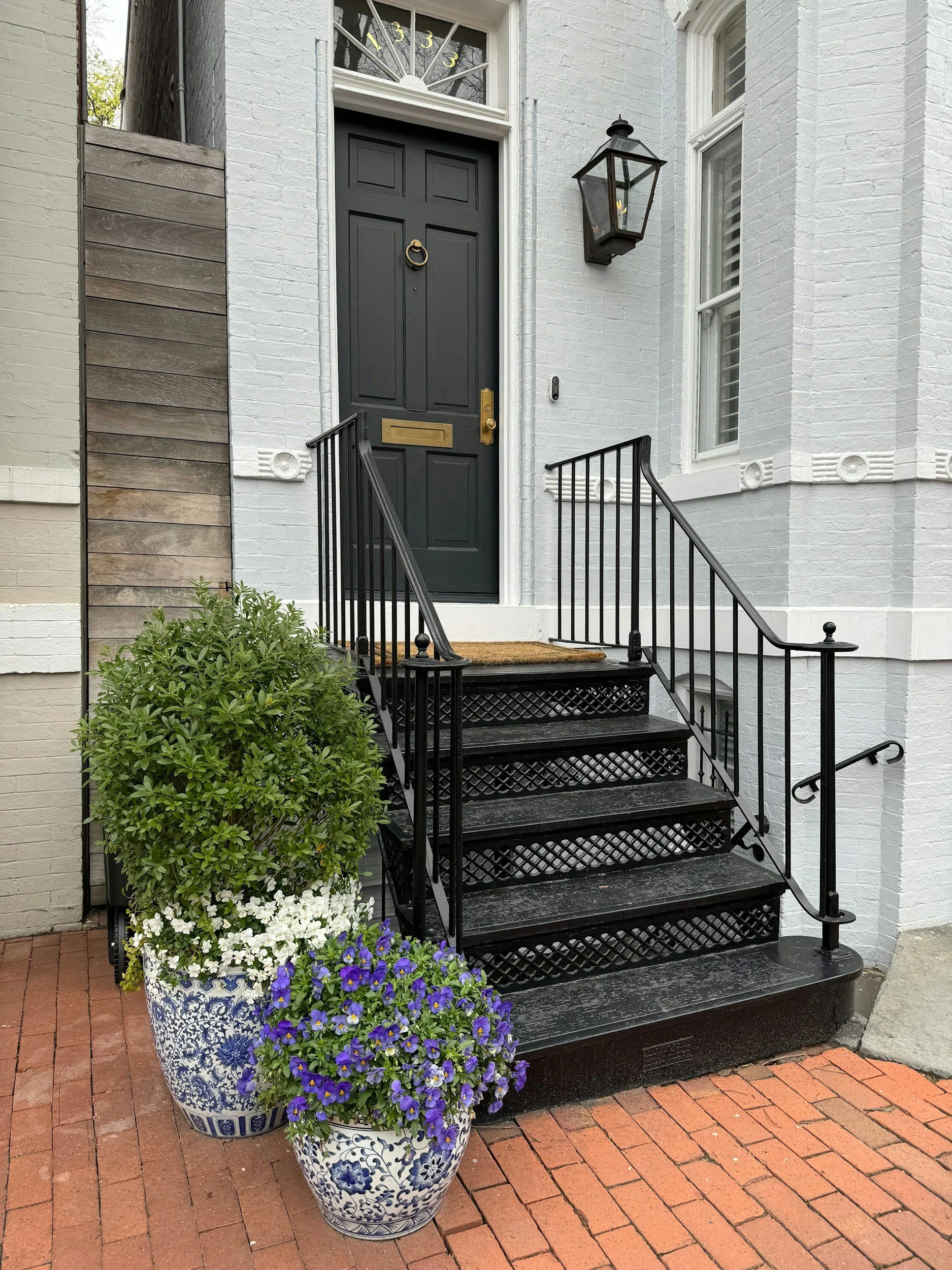 Front entrance of a house with black door, black metal steps, and white brick exterior. Two decorative flower pots with green and purple flowers are on the brick sidewalk. There is a black lantern light fixture beside the door and a doormat on the top step.