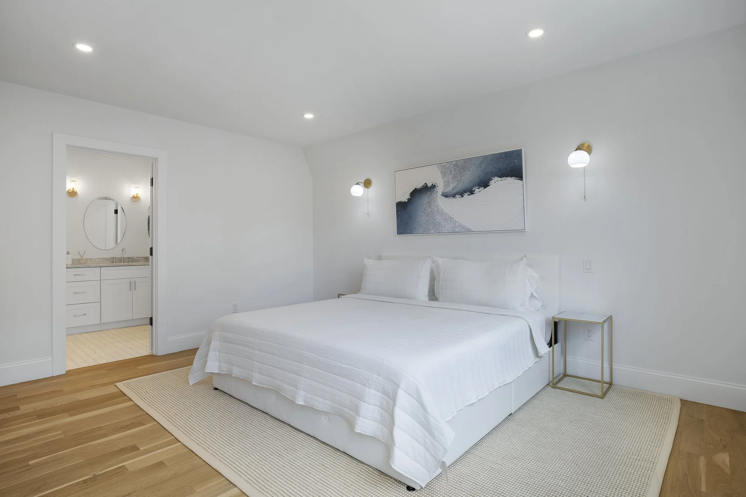Minimalist bedroom with white walls, bedding, and decor, wooden floor, and an attached bathroom visible through an open door.