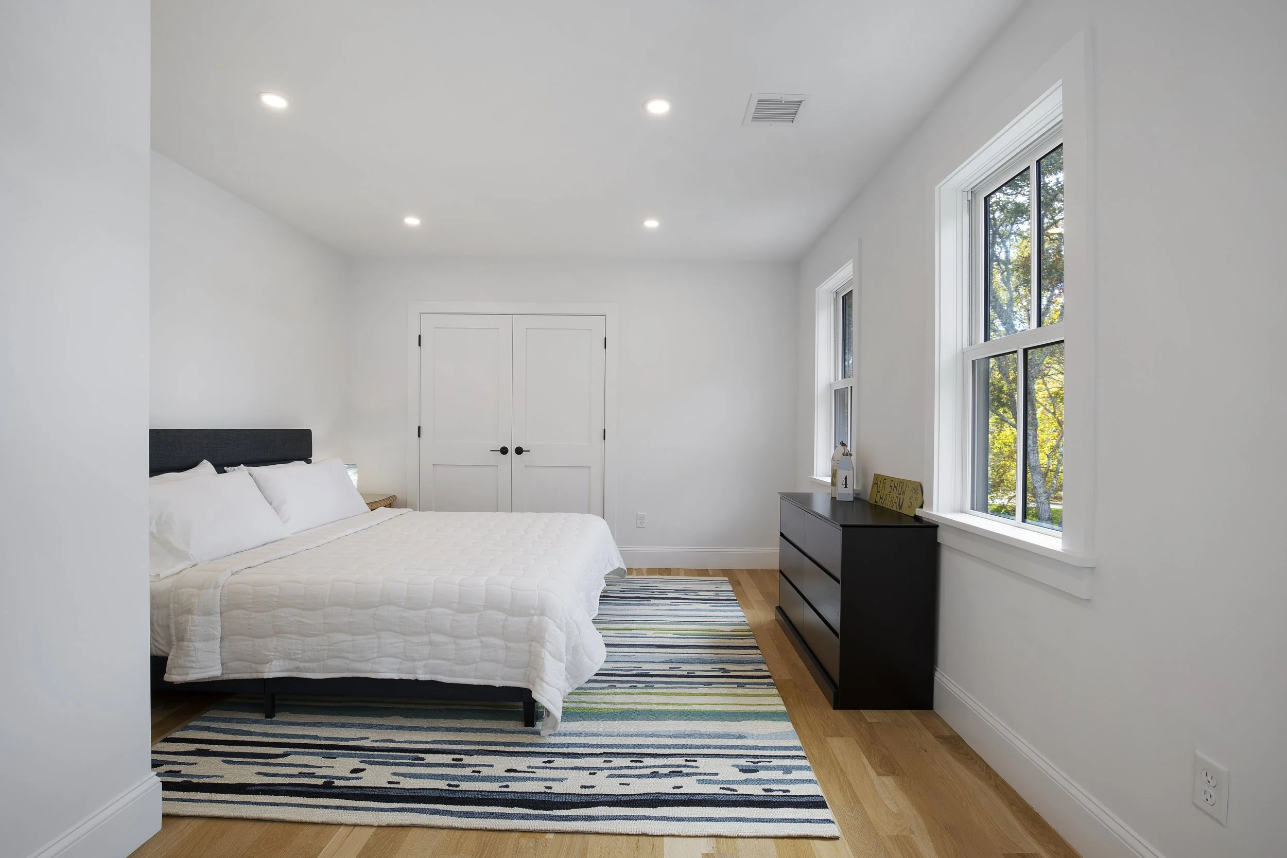Minimalist bedroom with white walls, hardwood floors, a bed with white bedding, a black dresser, and large windows showing trees outside.