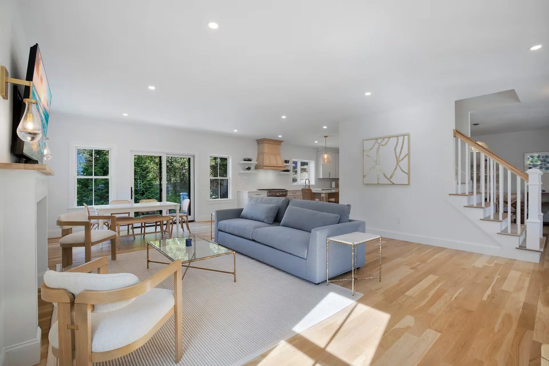 Bright open living room with gray sofa, white walls, light wood flooring, and large windows showing greenery outside.
