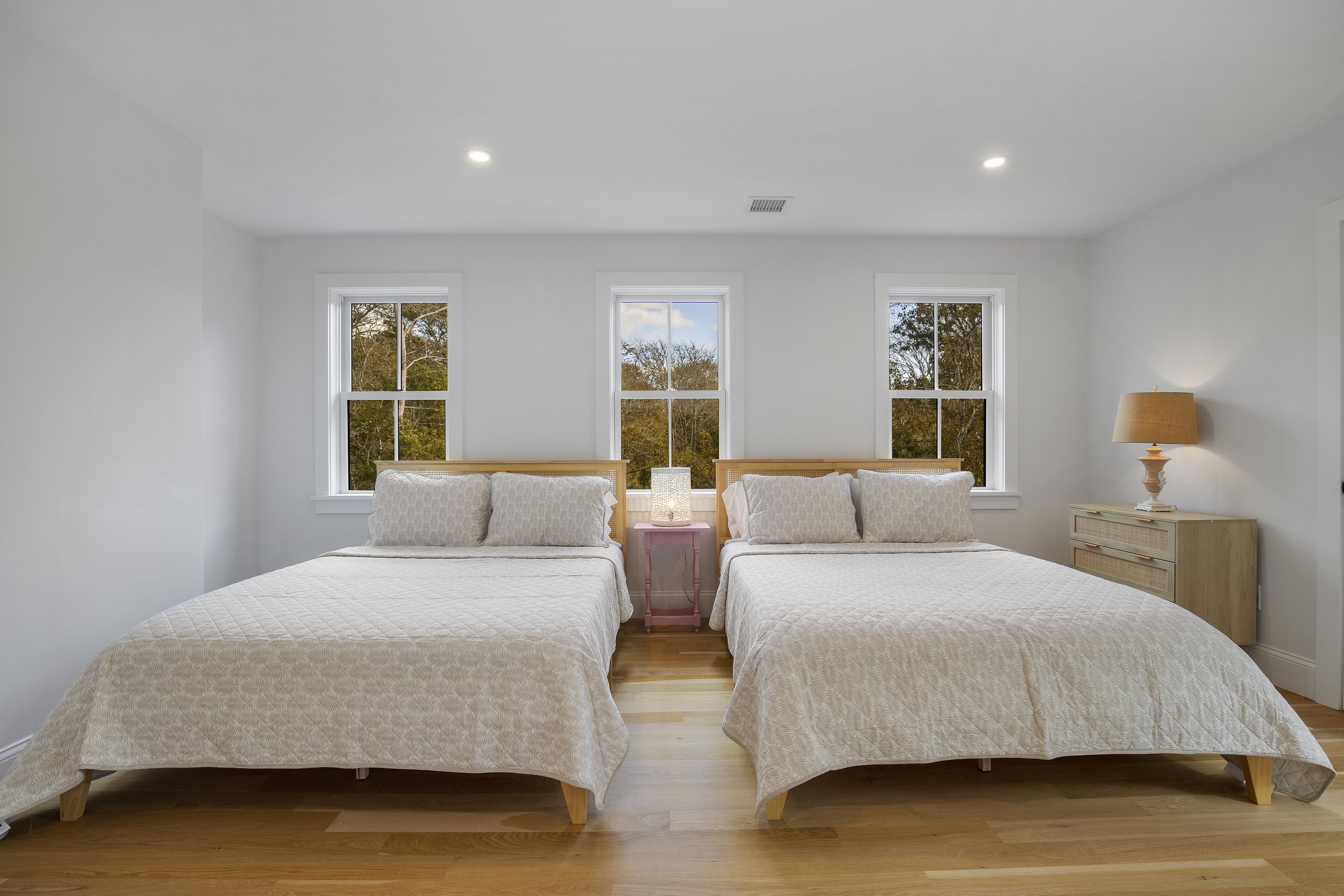 A bedroom with two beds with white bedding and pillows, a pink side table with a lamp between them, three windows showing trees outside, and a wooden dresser with a lamp on it, set in a bright room with hardwood floors.