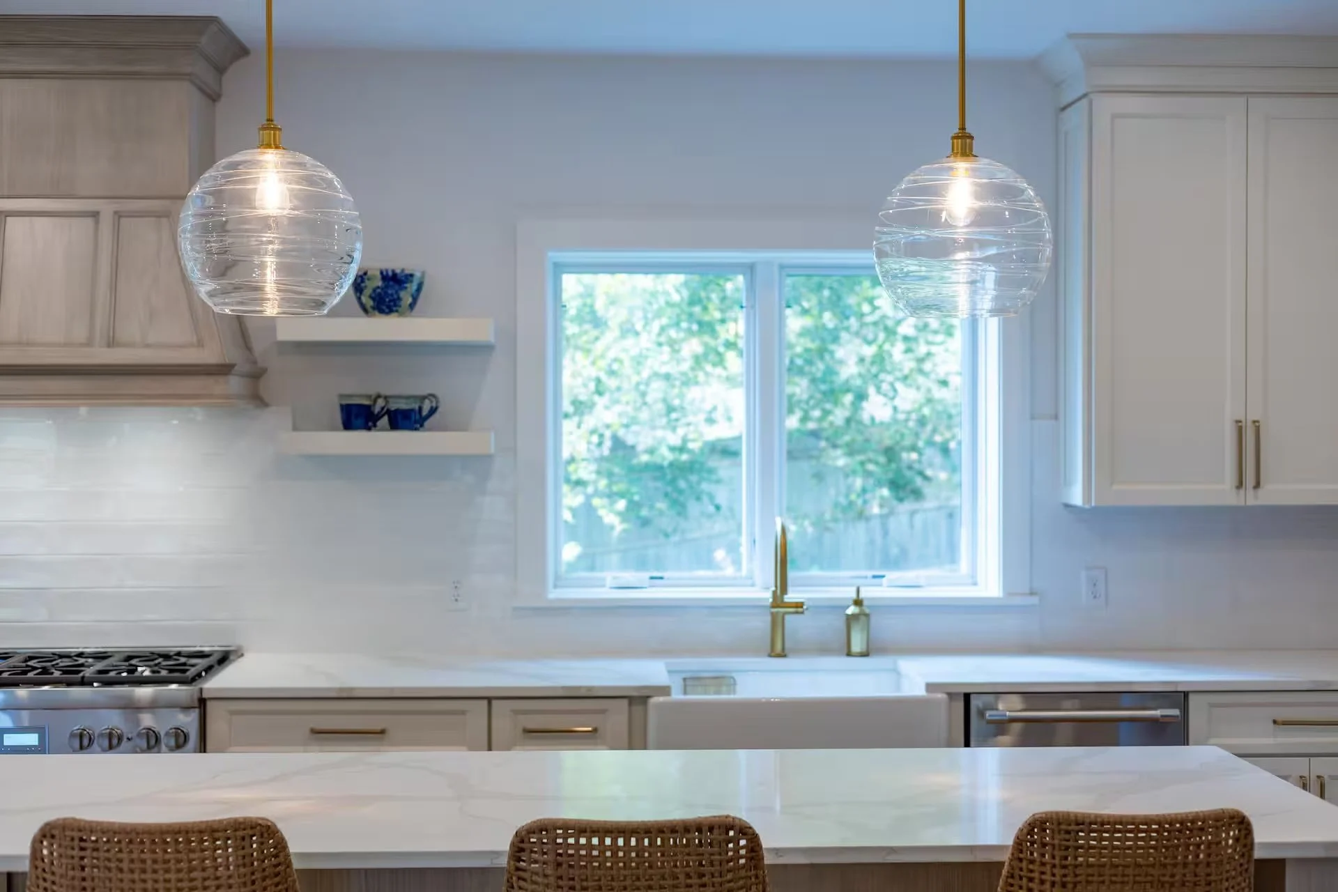 Modern kitchen with white cabinets, a large window, and two hanging pendant lights with glass globes. There is a white countertop and chairs at a kitchen island.