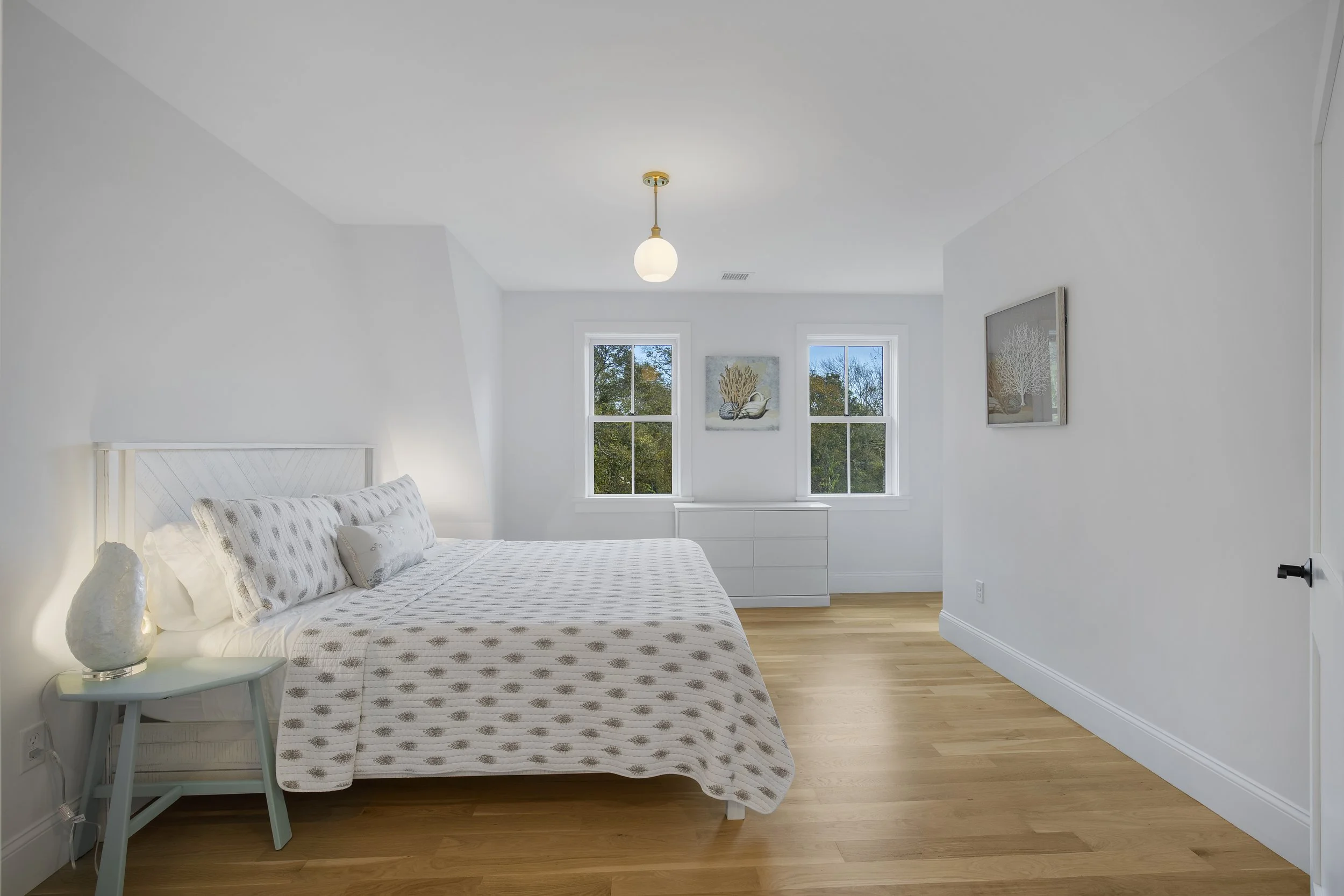 Bright bedroom with a white bed, patterned bedding, a nightstand with a lamp, and two windows with outside greenery.