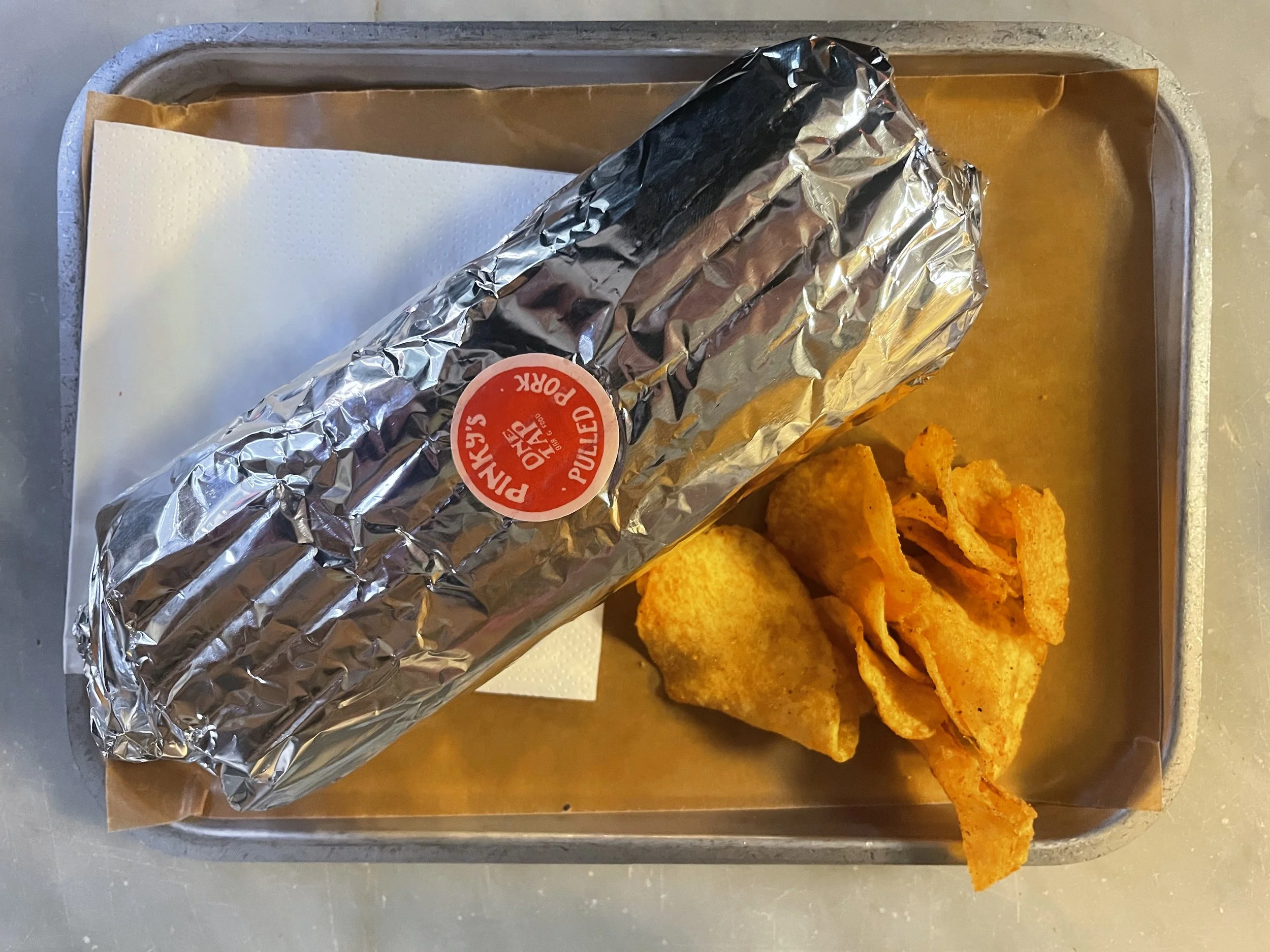A tray with a piece of foil-wrapped cooked chicken with a round red label that says "Peters Organic Fully Cooked Rotisserie Chicken" and a serving of seasoned potato chips.