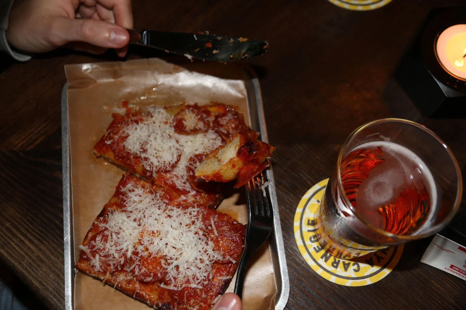 Close-up of three slices of pepperoni pizza topped with shredded cheese, on a metal tray lined with parchment paper, with a hand holding a fork. To the right, there is a glass of beer with froth on top, set on a yellow and black branded coaster on a dark wooden table, and a lit candle in a black holder.