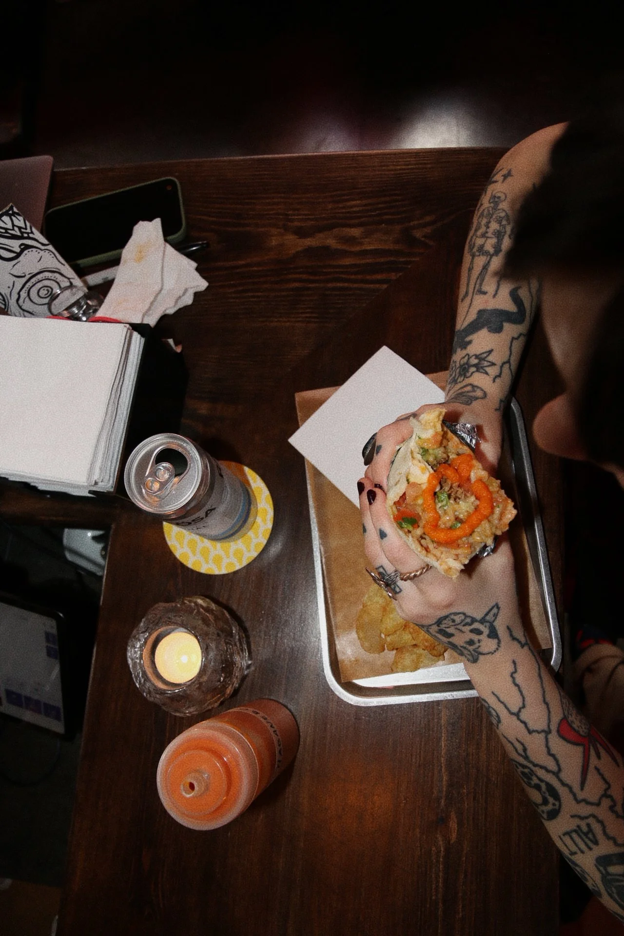 Person holding a slice of sushi with tattoos on their arm, on a dark wooden table with a candle, drinks, and papers.