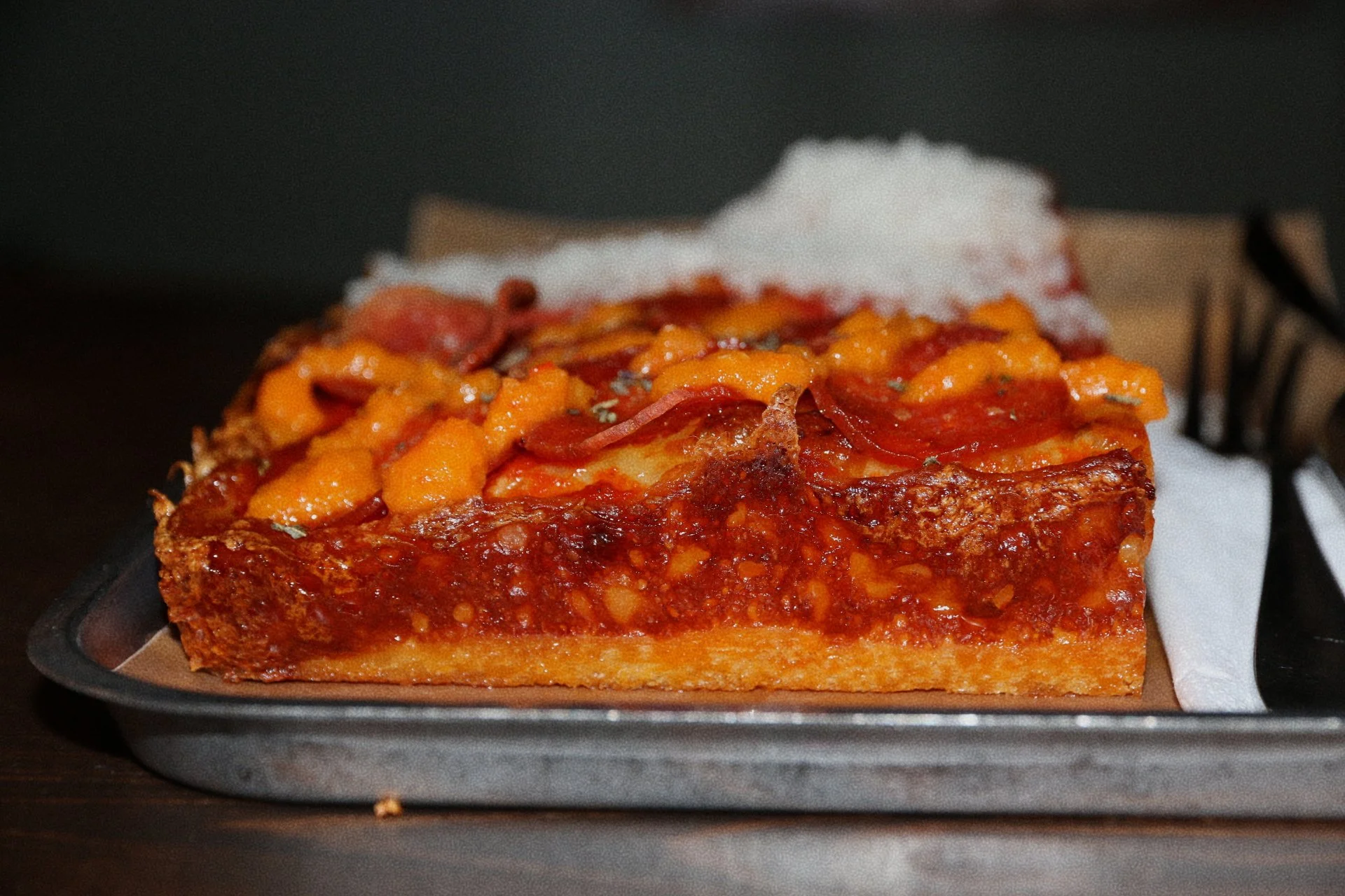 A slice of pepperoni pizza with cheese and tomato sauce on a black tray, with a napkin and a fork in the background.