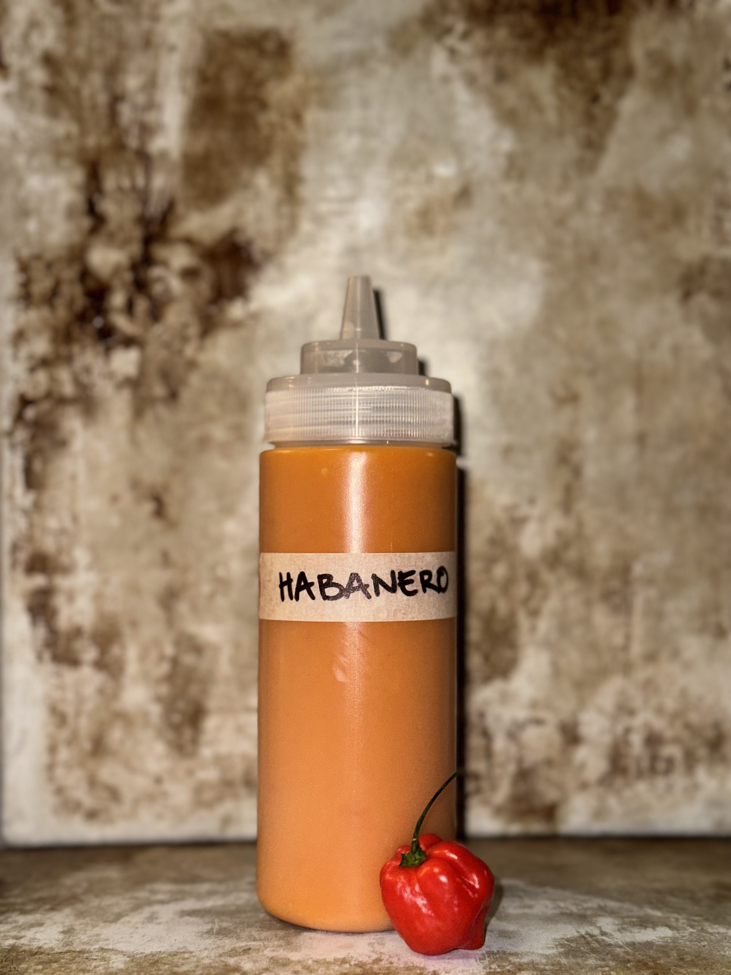 A peach-colored bottle with a label that says 'HABANERO' in black handwriting, placed on a textured surface with a red habanero pepper in front of it, and a blurred stone background.
