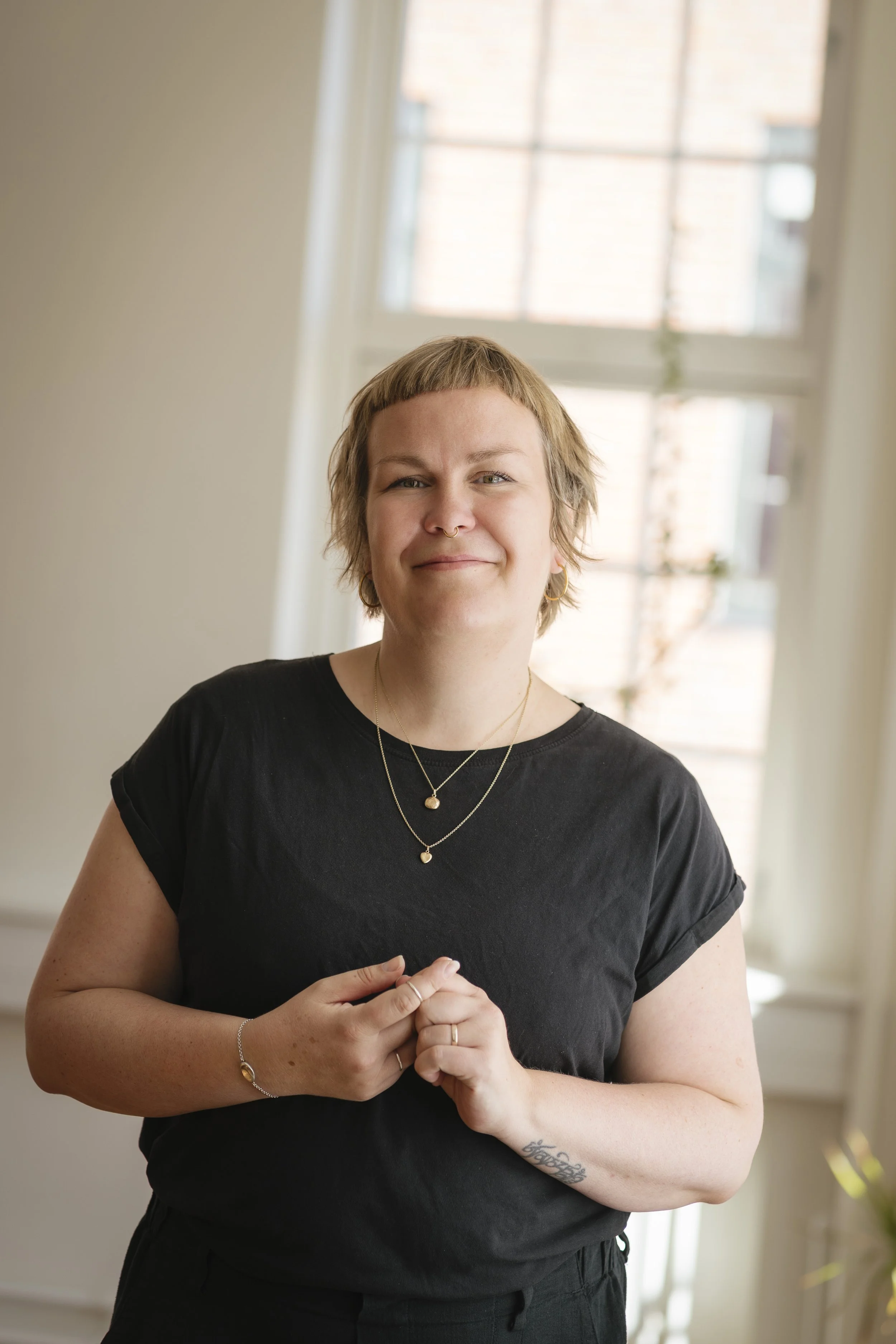 En ung kvinde med kort, lyst hår, iført sort T-shirt og smykker står indendørs foran et stort vindue, der slipper masser af naturligt lys ind.