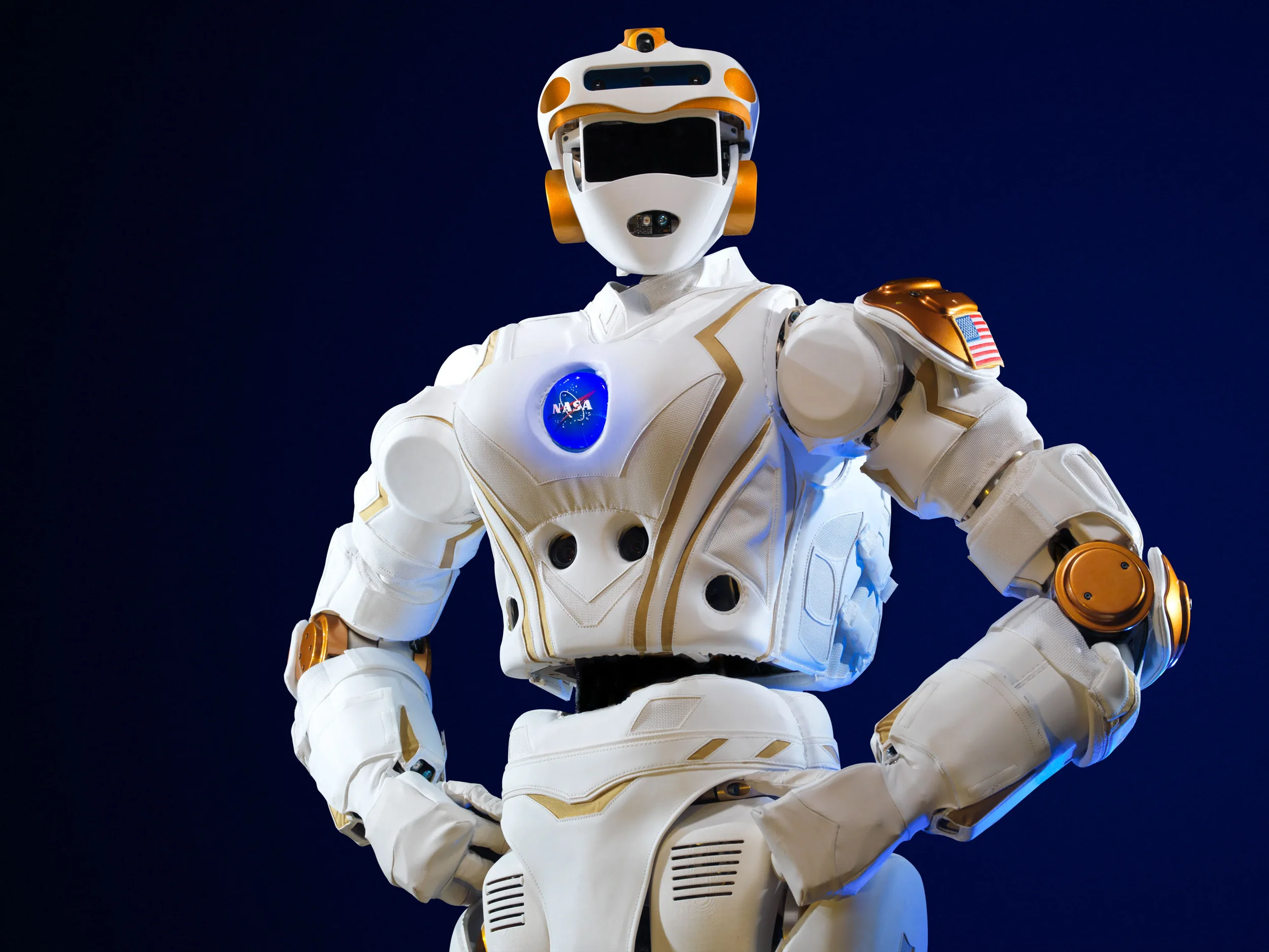A humanoid robot with a white and gold armor-like exterior, featuring the NASA logo on its chest, set against a dark blue background. The robot has a helmet with a visor and hoses or cables connecting various parts, and an American flag patch on its shoulder.