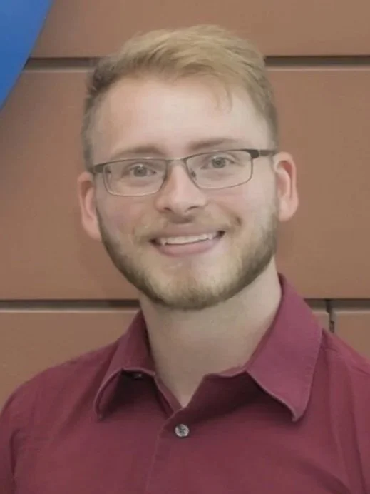 A young man with glasses, short light brown hair and a beard, smiling in front of a brown wooden wall, wearing a maroon shirt.