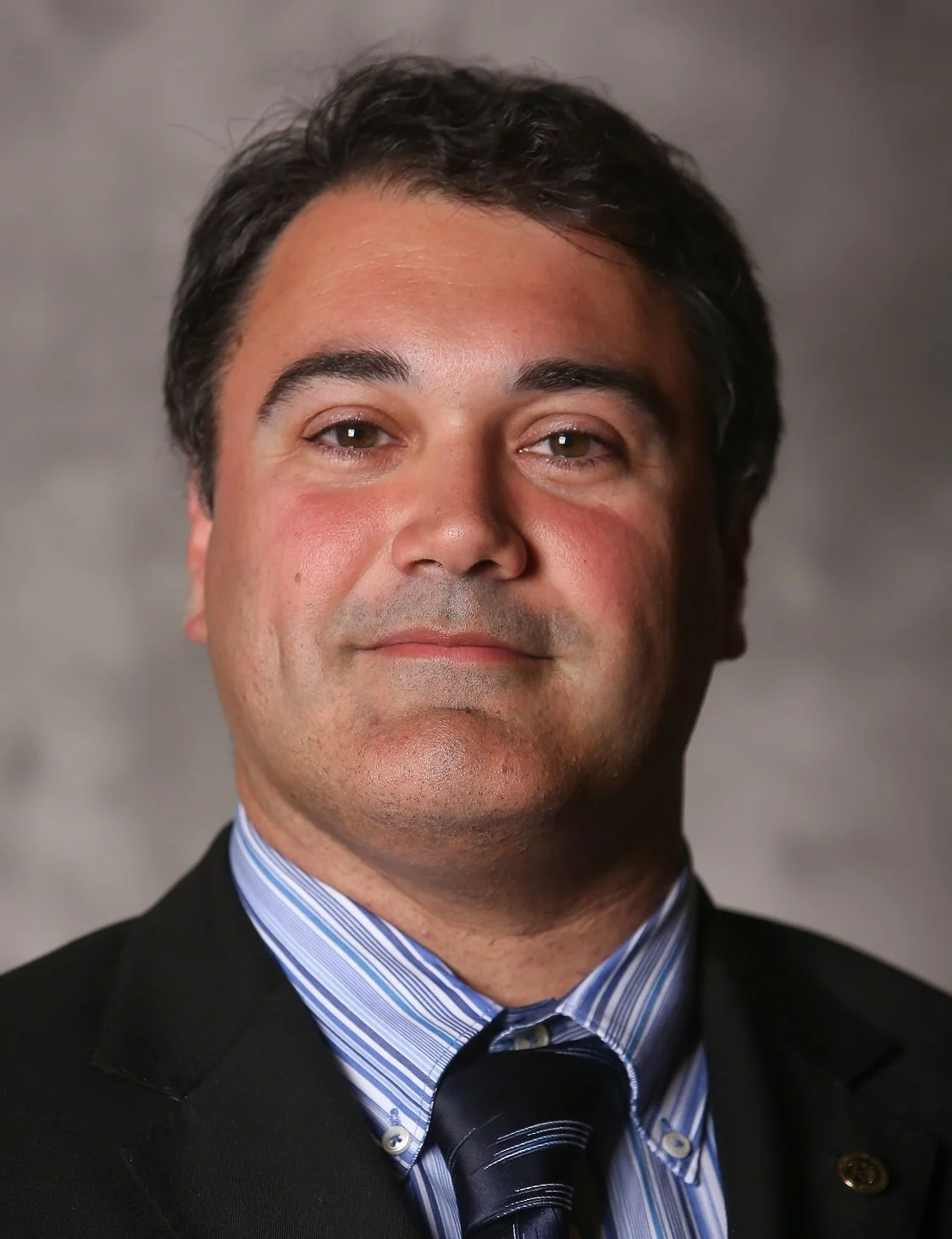 Portrait of a man wearing a black suit, striped blue shirt, and a striped tie, with a neutral background.