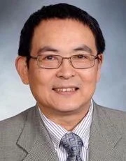 A professional headshot of an Asian man with short black hair and glasses, wearing a gray suit, white shirt, and a patterned tie, smiling against a bluish background.
