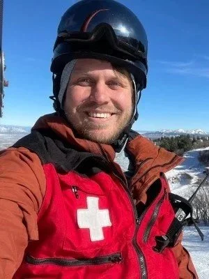 A smiling man wearing a black helmet and a red jacket with a white cross emblem, in a snowy outdoor setting under a clear blue sky.
