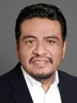 Headshot of a man with dark hair, beard, and mustache, wearing a dark blazer and white shirt, against a gray background.