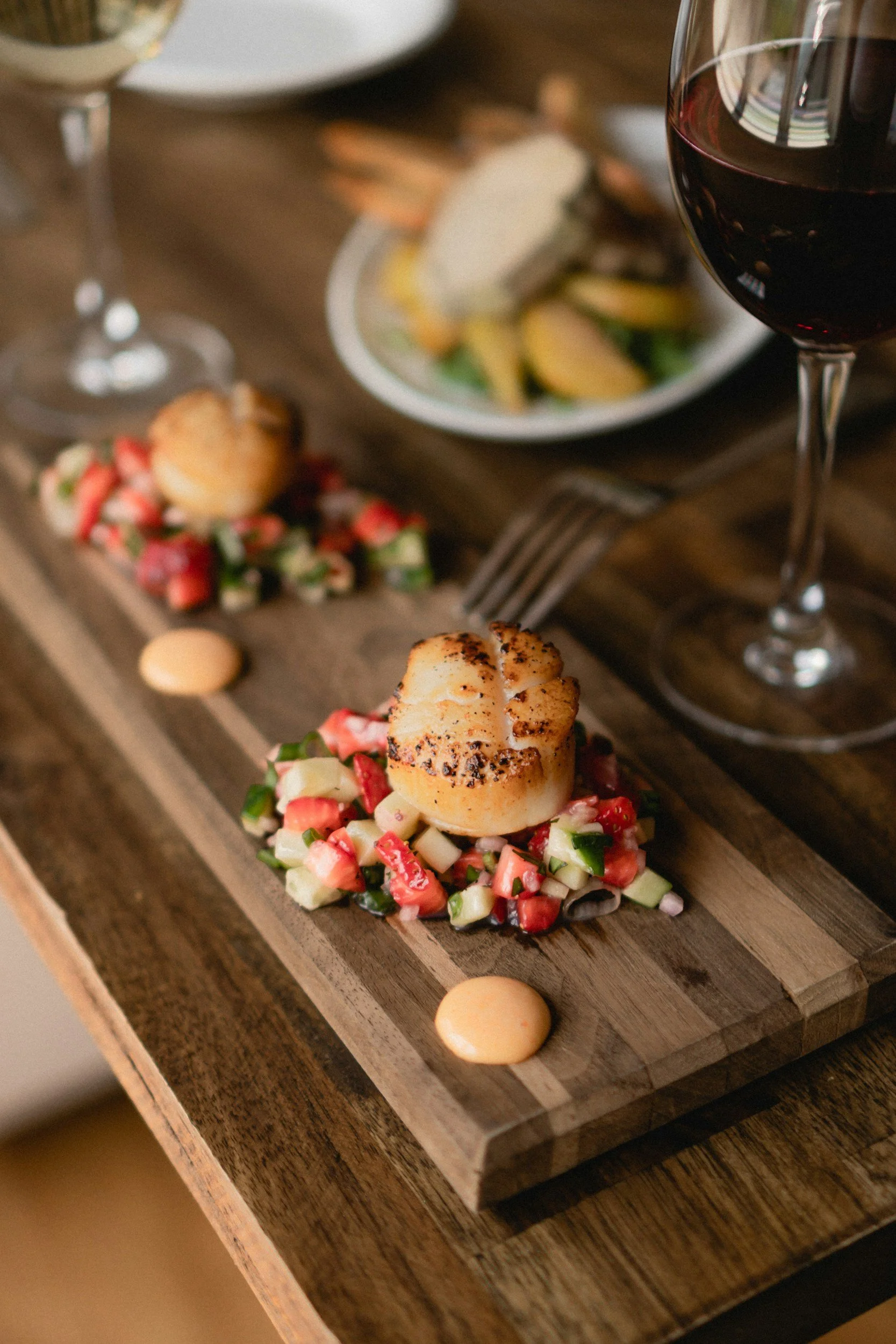 A wooden serving board with a scallop topped with a diced vegetable salad, garnished with small dollops of sauce and surrounded by chopped vegetables and two glasses of red wine.