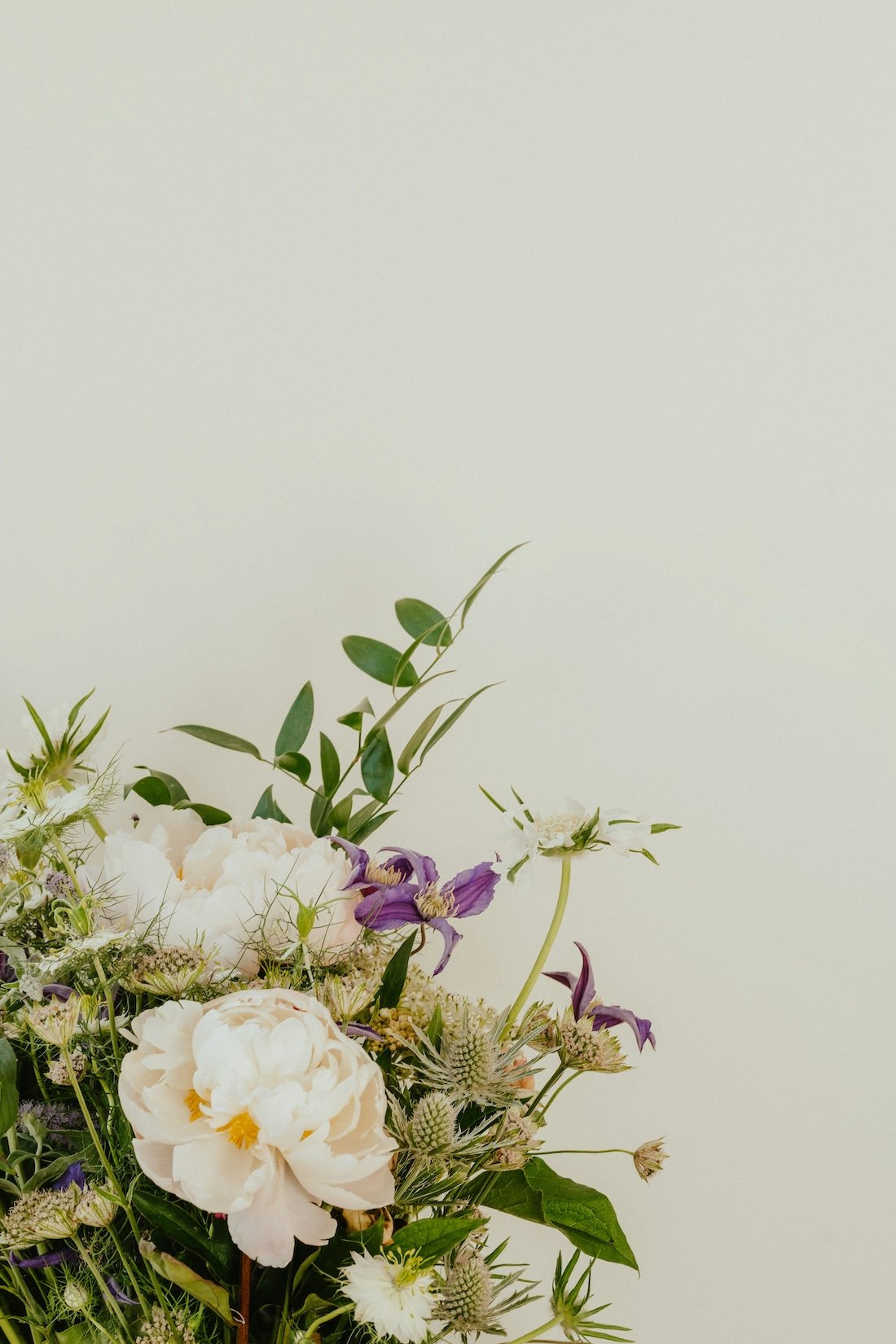 A bouquet of white peonies, purple irises, and various greenery set against a plain, light-colored background.