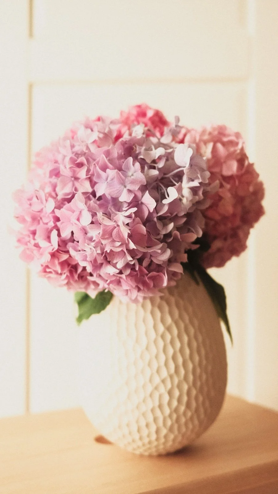 Pink and purple hydrangea flowers in a textured cream-colored vase on a light wood surface.