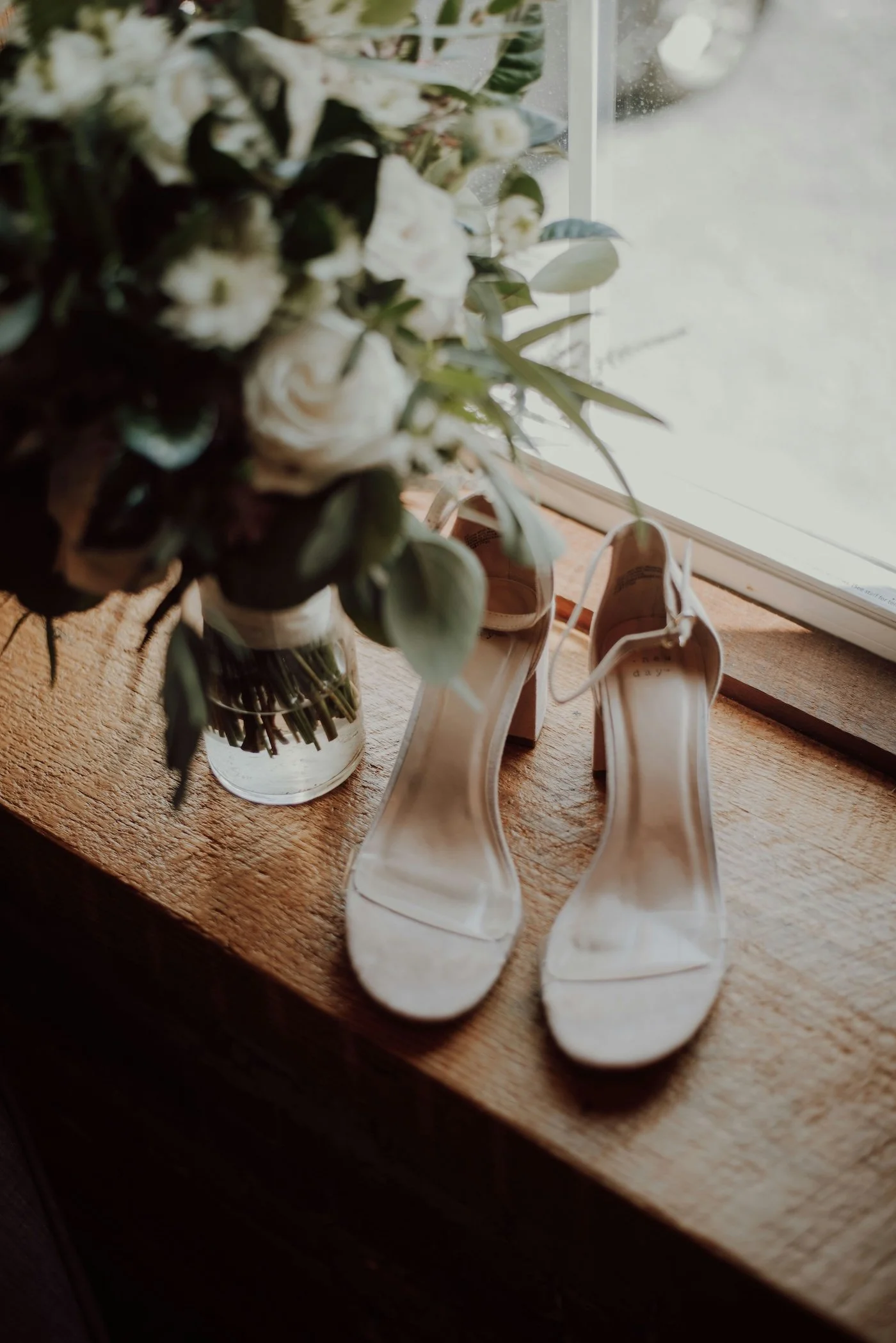 A pair of beige high-heeled shoes on a wooden surface next to a vase of white flowers near a window.
