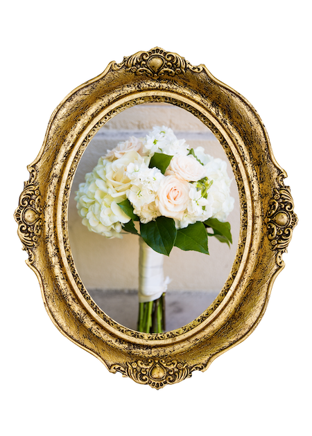 A bouquet of white roses and flowers with green leaves, reflected in an ornate gold oval mirror.