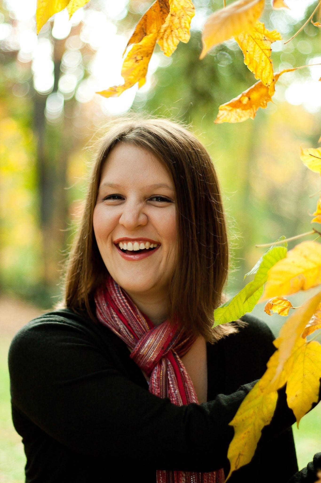 A woman with shoulder-length brown hair smiling outdoors during fall, surrounded by yellow and orange leaves.