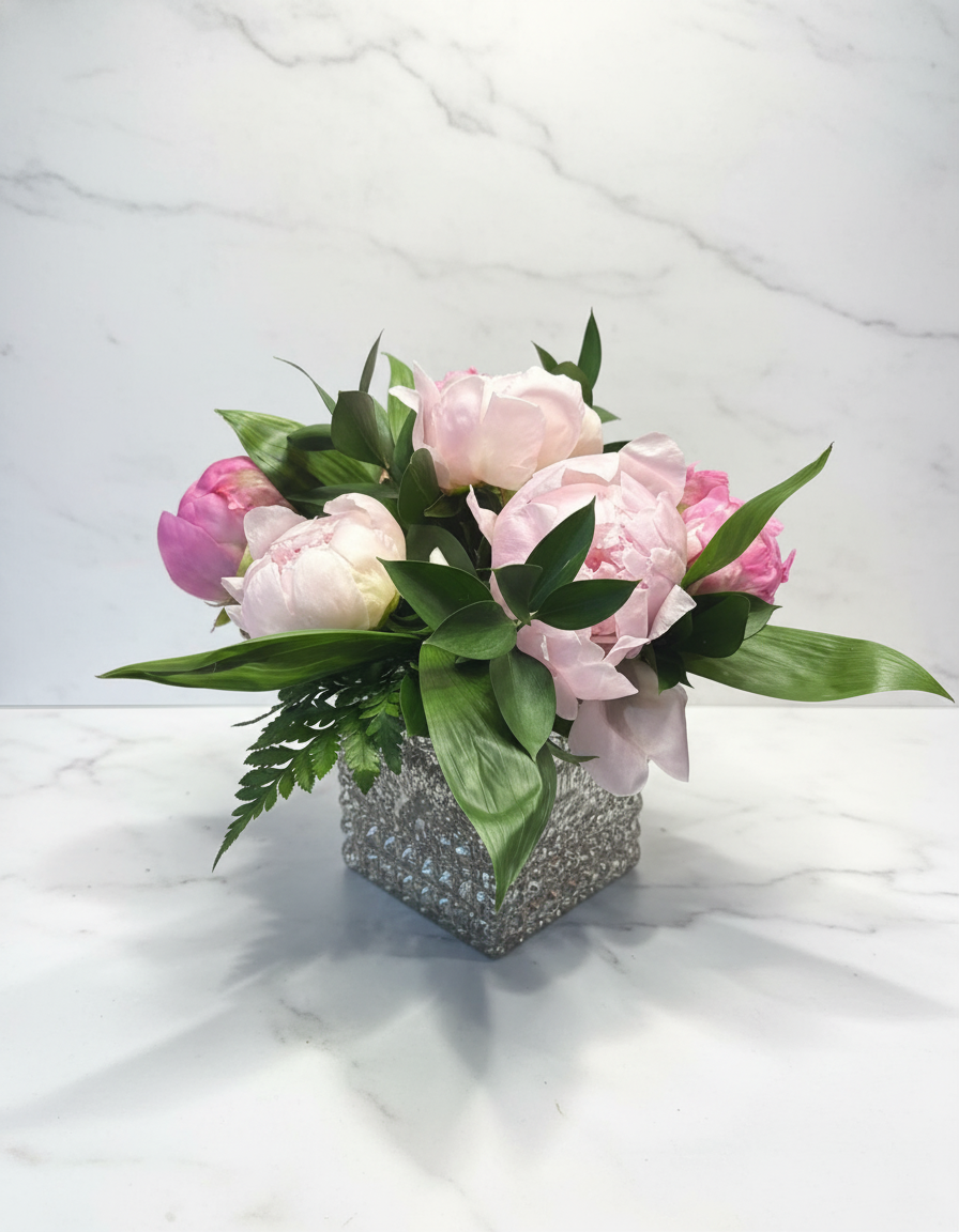 A bouquet of light pink and white peonies with green leaves in a textured, square vase on a white marble surface against a white marble wall background.