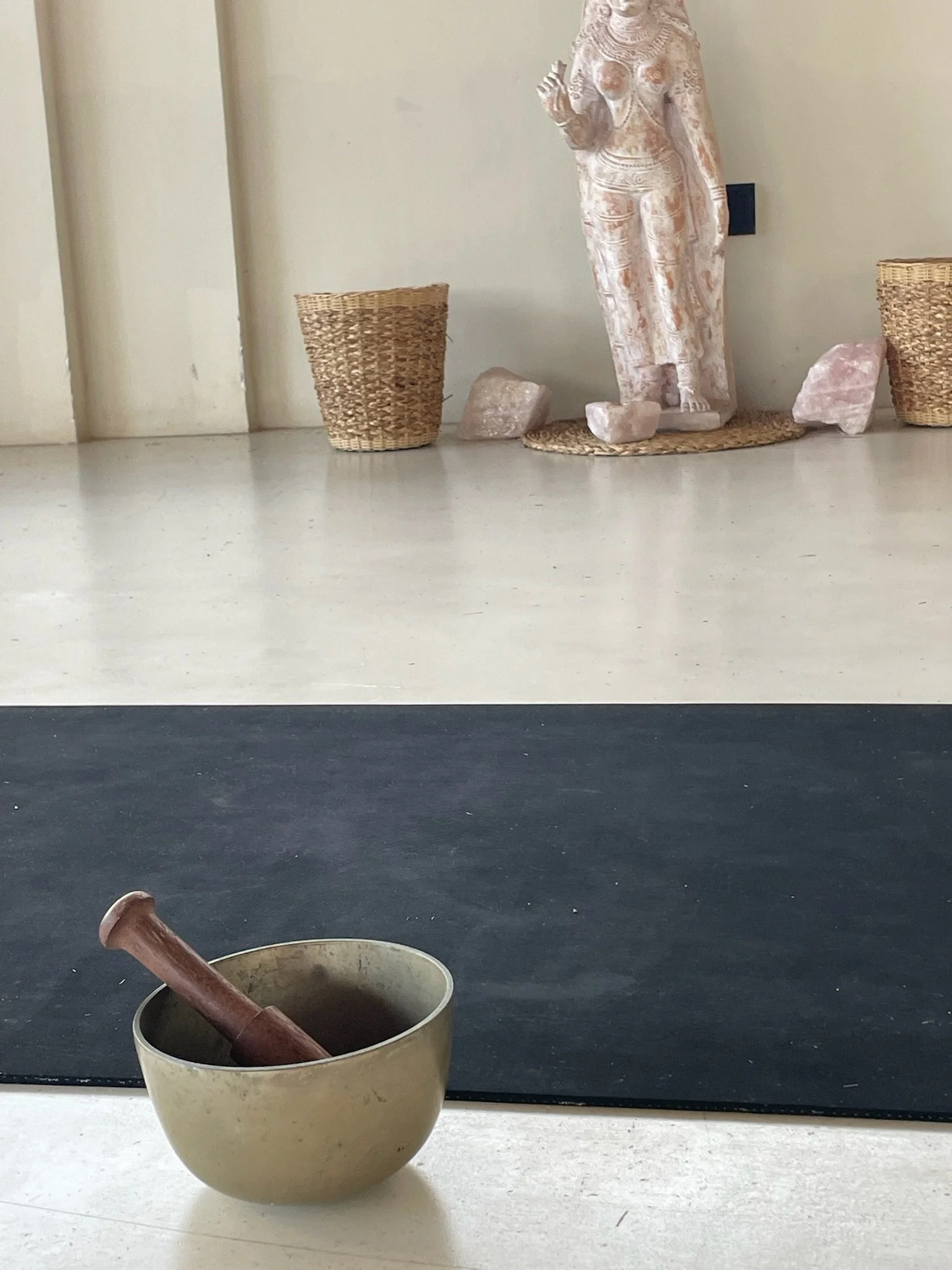 A beige ceramic pot with a wooden pestle inside, placed on a light-colored surface. Behind it, a black and cream striped background, and to the right, a round woven basket, a pink Himalayan salt block, and a white sculpture of a woman holding a small object.