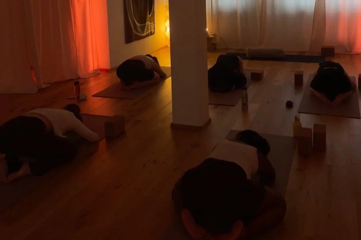 People practicing yoga in a room with warm lighting, sitting on yoga mats in a kneeling position.