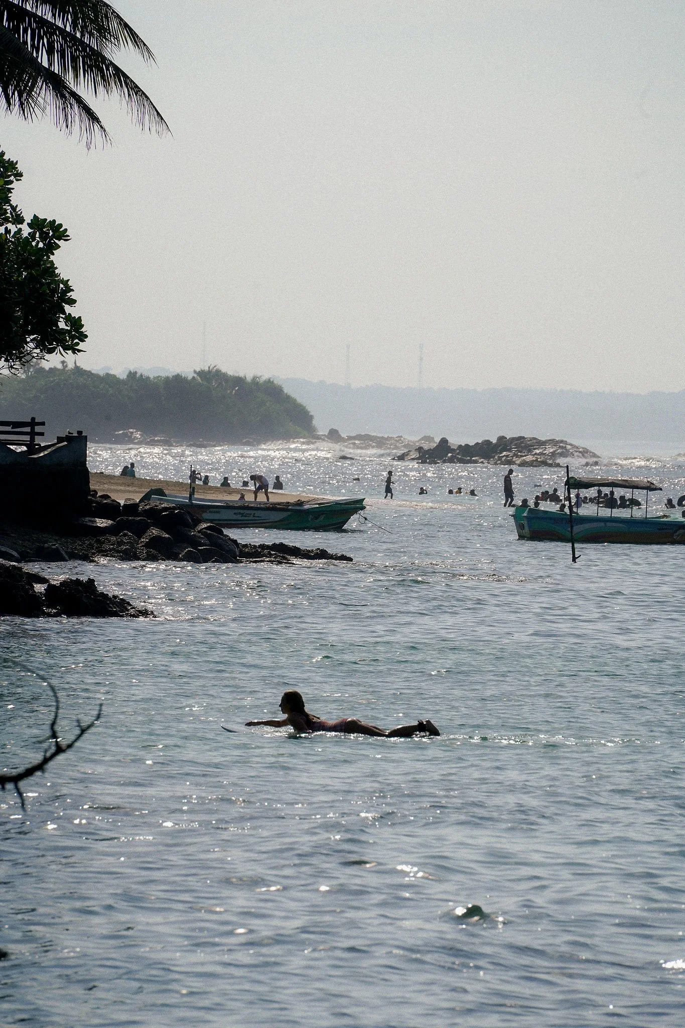 A beach scene with boats, people swimming and playing in the water, and trees along the shoreline, with hills and communication towers in the background.