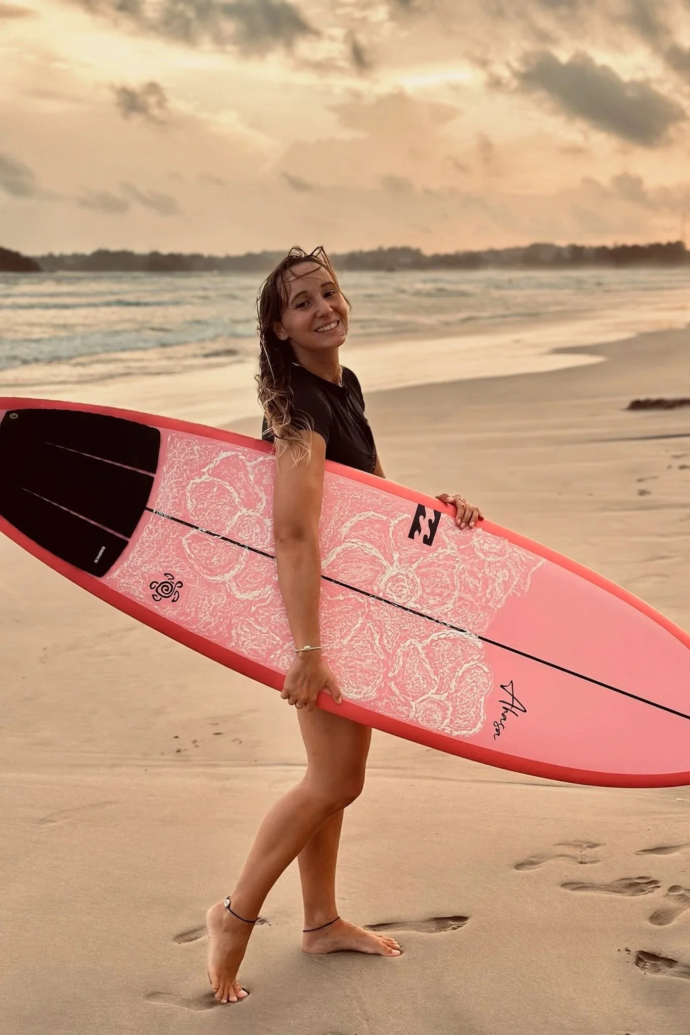 A woman smiling and holding a pink surfboard on the beach at sunset, with footprints in the sand and waves in the background.