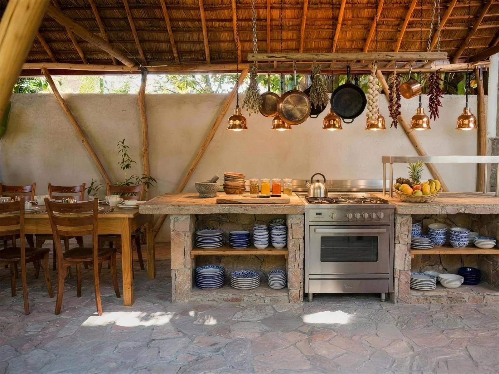 An outdoor kitchen with a rustic design. It features a stone and wood counter, a stainless steel stove, shelves with blue and white dishes, hanging pots and dried herbs, a bowl of assorted fruits, and a dining table with chairs.