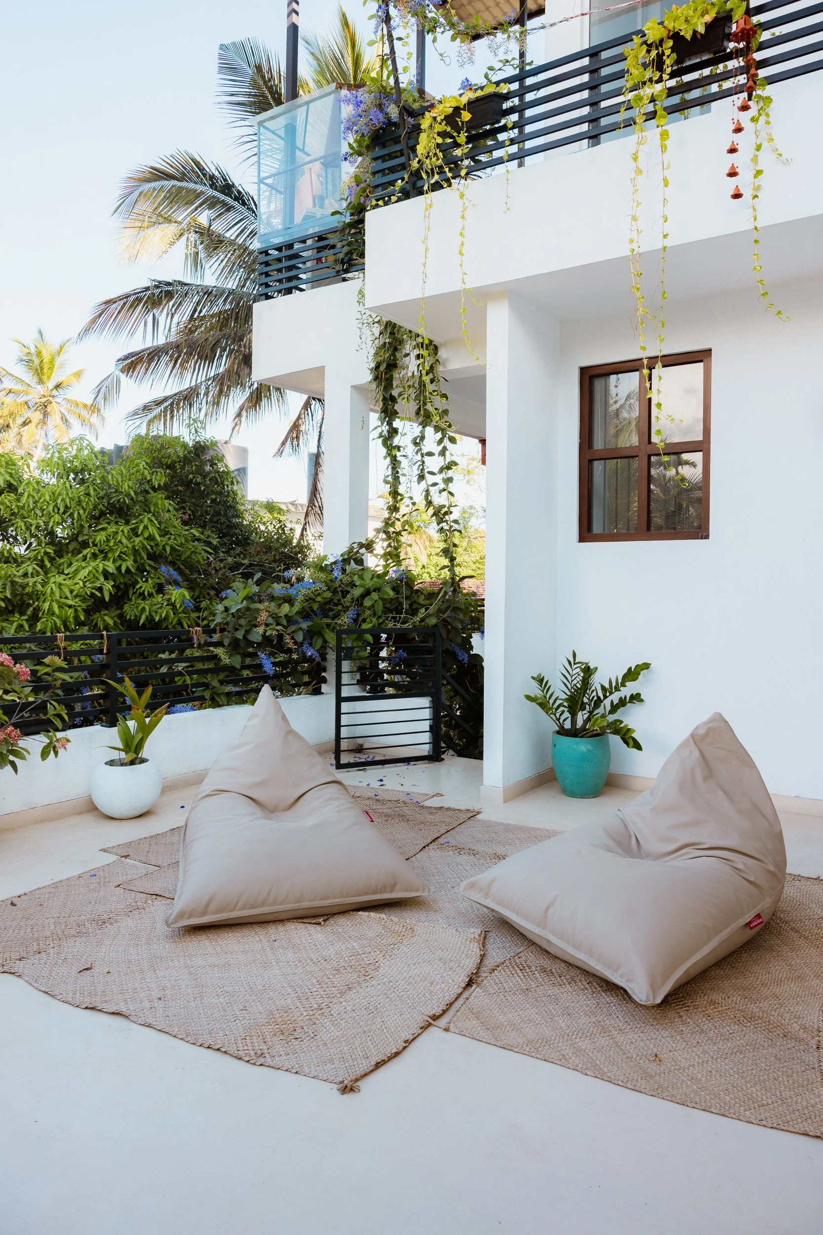 A balcony with green plants and hanging vines, and a white wall with a small window. The balcony has a glass railing and outdoor furniture, including bean bag chairs on a woven rug.