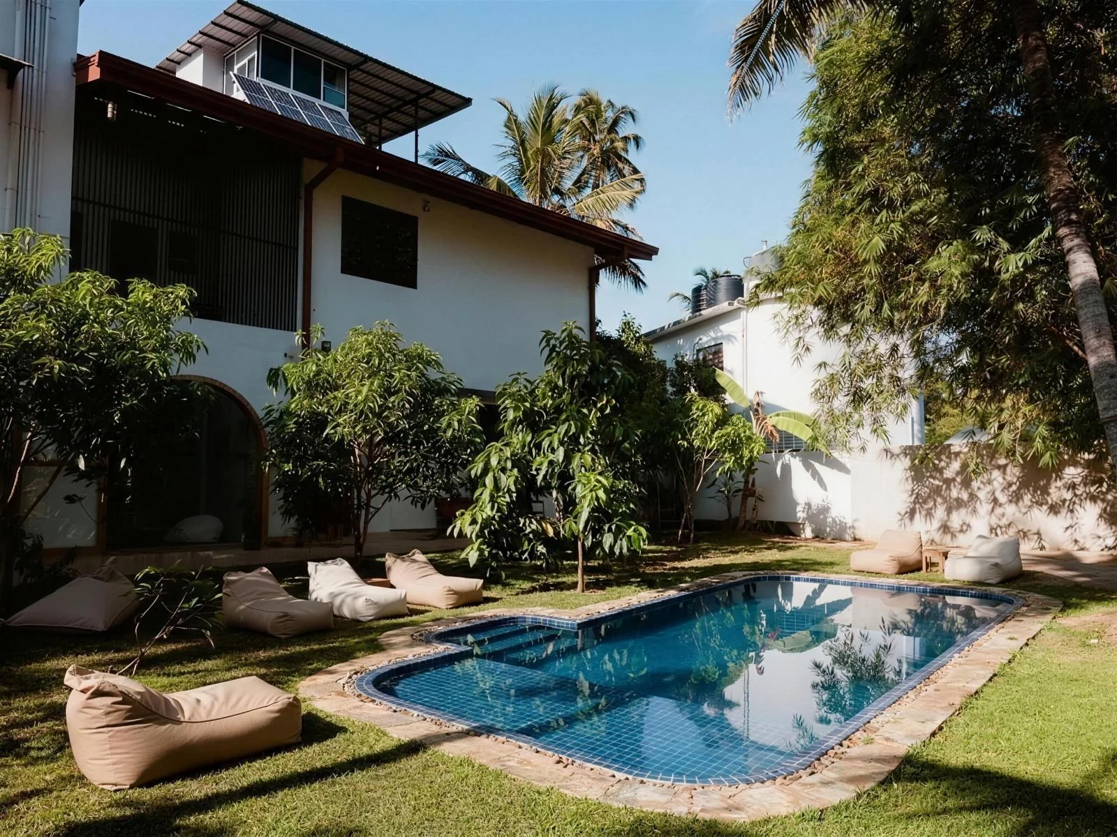 A backyard with a small swimming pool, surrounded by lounge chairs and lush green trees, with a white house featuring large windows and solar panels on the roof in the background under a clear blue sky.