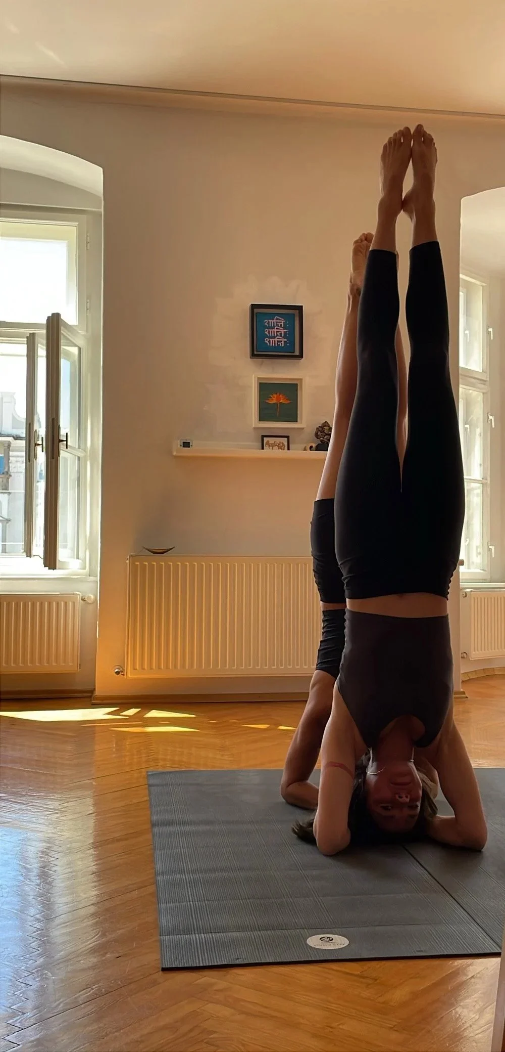 A woman practicing yoga in a headstand pose indoors on a gray yoga mat. She is wearing black leggings and a black top, with windows and artwork on the wall in the background.