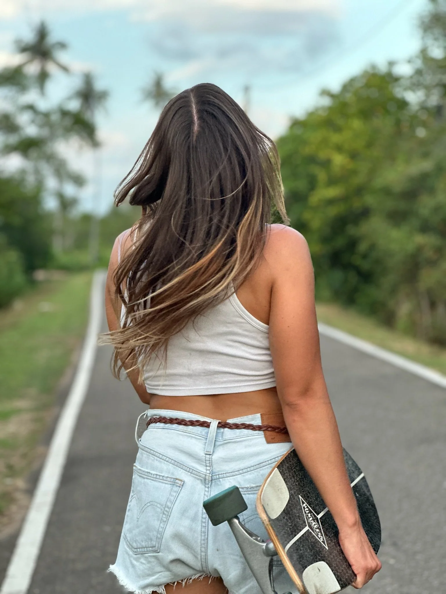 A woman with long, wavy hair wearing a white tank top and distressed denim shorts, holding a skateboard and another skateboard hanging from her hand on a paved road with greenery and palm trees in the background.