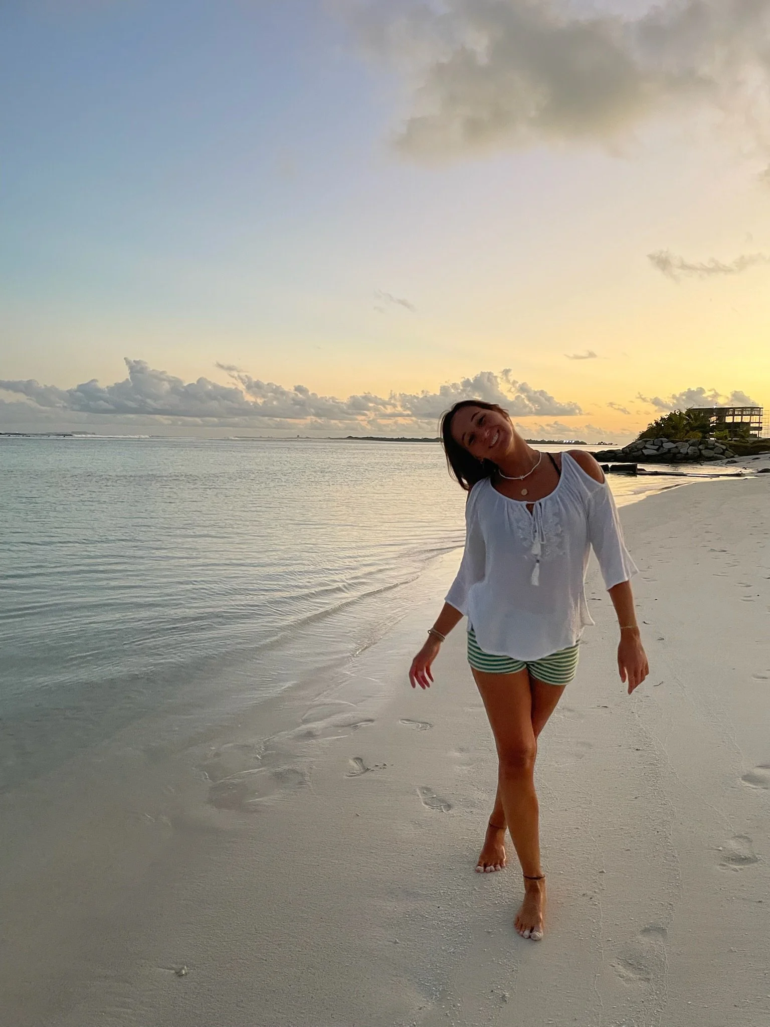A woman smiling on a beach at sunset, wearing a white top and striped shorts, walking barefoot on the sand with the ocean and clouds in the background.