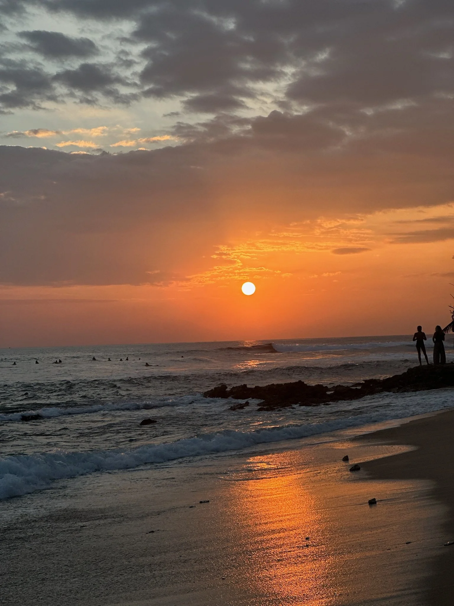 Sunset over the ocean with a partly cloudy sky, two people walking along the beach, and waves crashing onto the shore.