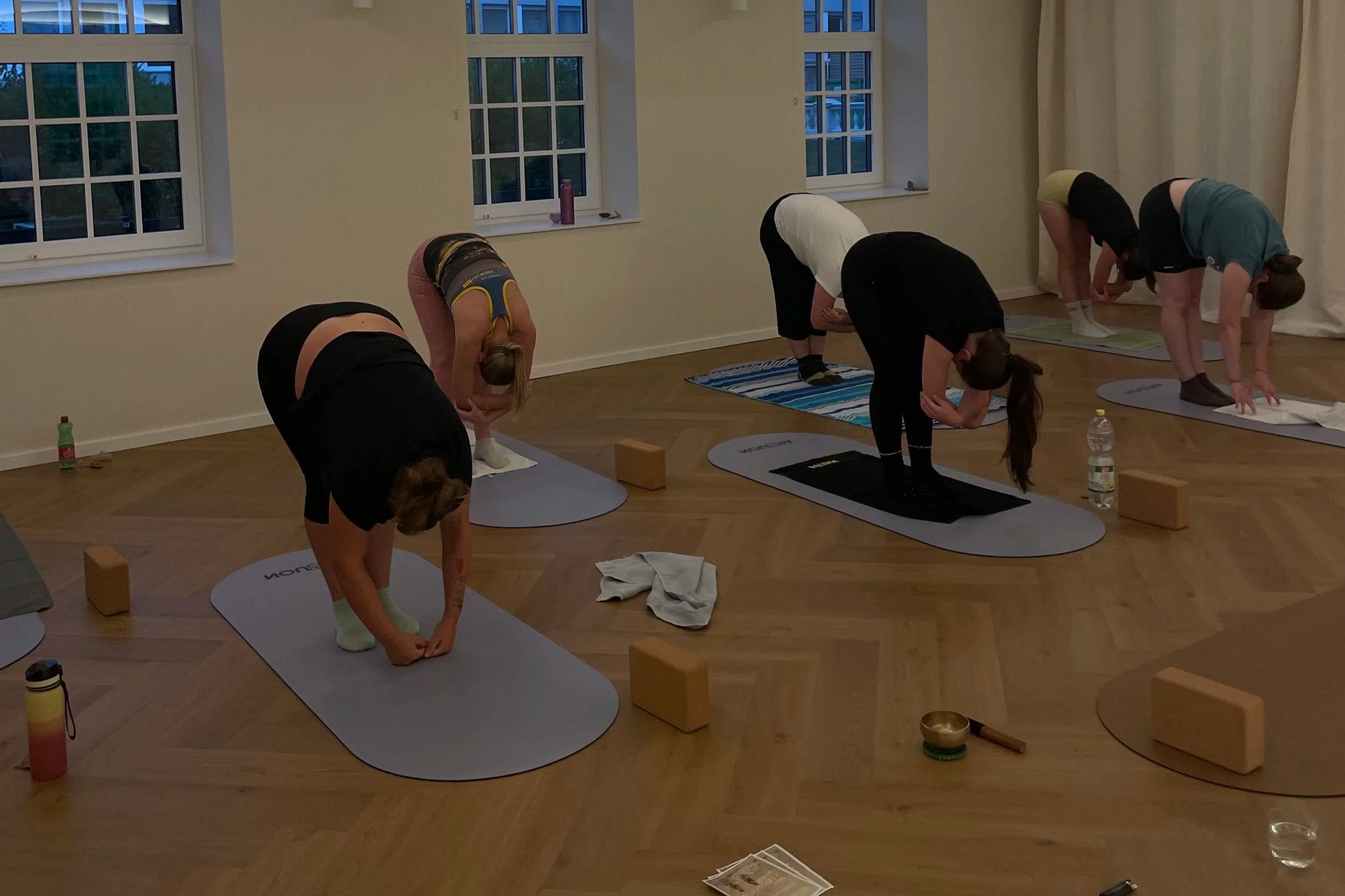 A group of five people practicing yoga in a room with wooden floors and large windows. They are in a forward bend pose on yoga mats, dressed in athletic clothing. Water bottles and yoga props are visible around them.