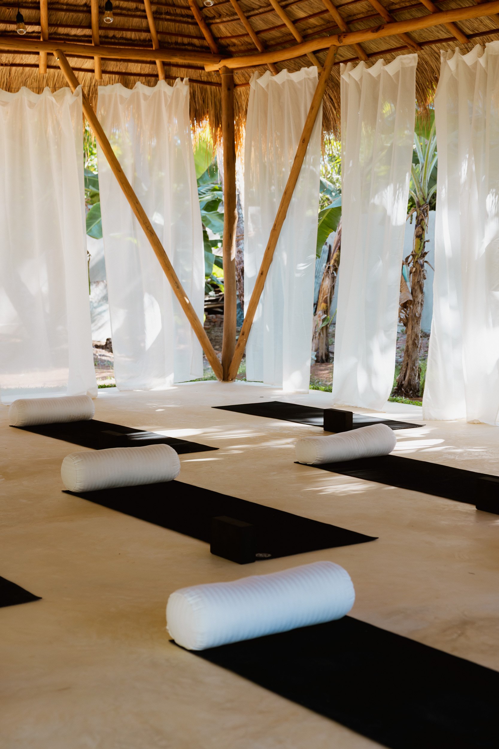 Indoor yoga space with black mats, white bolster pillows, and large windows with white curtains. Outside view includes tropical plants and trees, and the space has a thatched roof.
