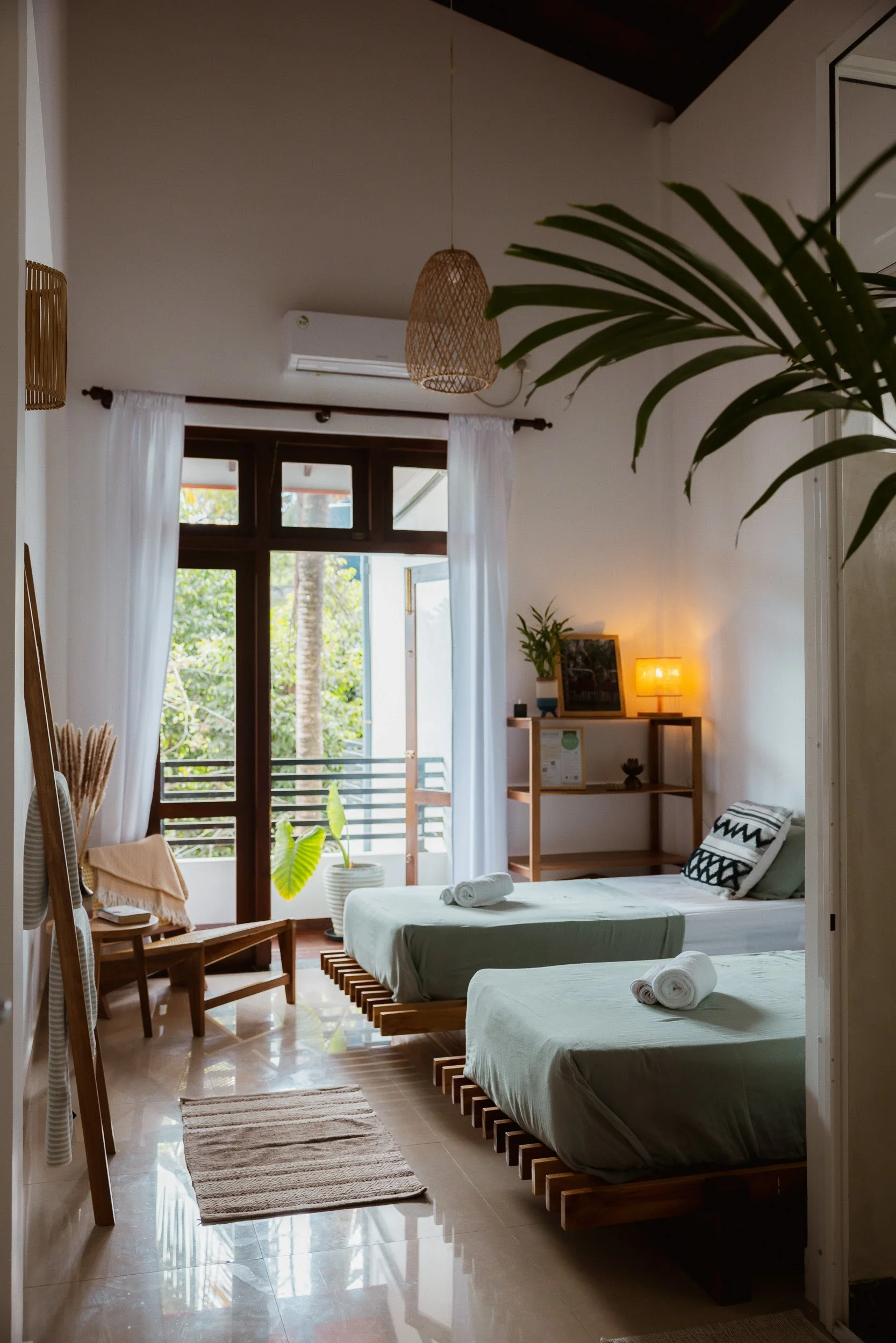 A cozy bedroom with two beds, each with rolled towels, near a large window with white curtains. There is a small wooden chair with a beige blanket, a potted plant, and a shelf with a lamp and framed picture. The room features a natural, minimalist decor with wooden furniture and soft lighting.