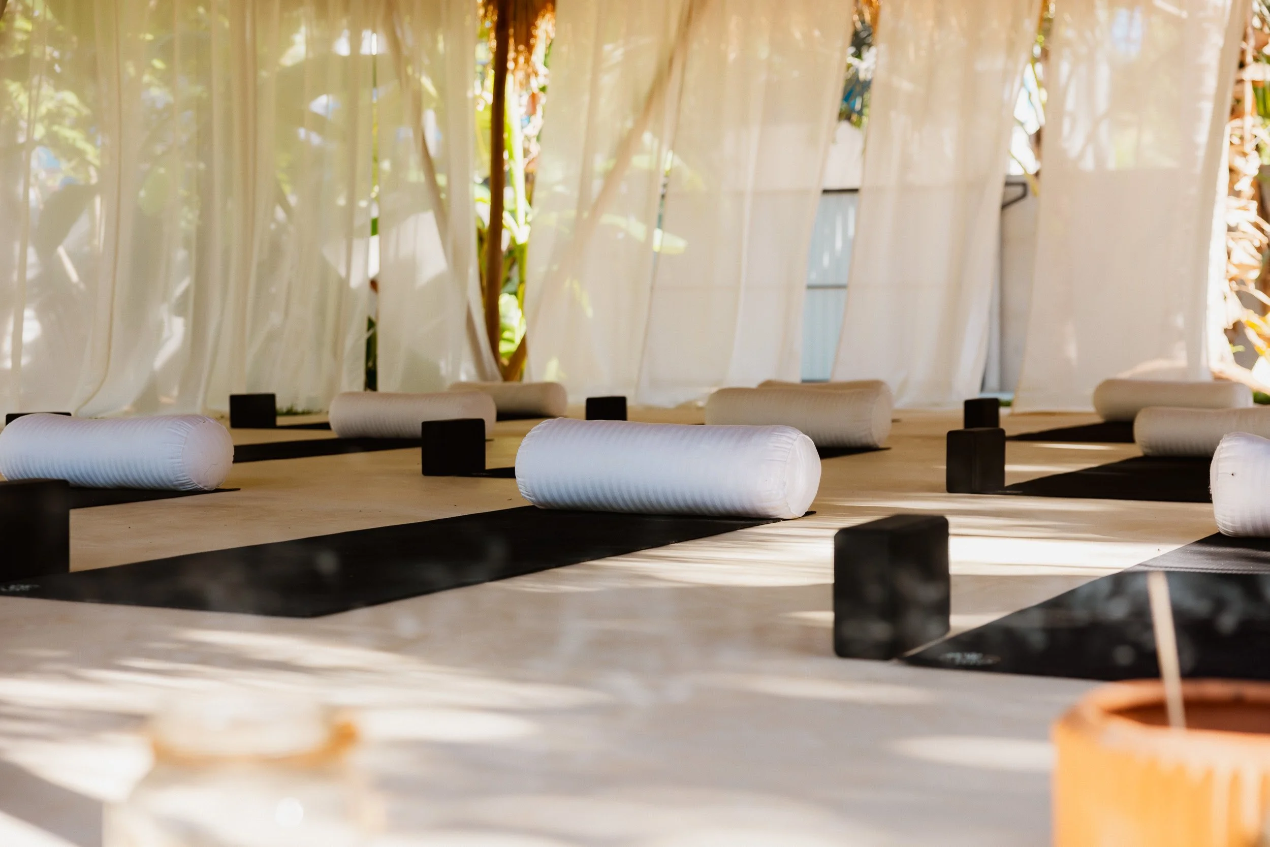 Indoor yoga and meditation space with white bolsters, black mats, and linen curtains allowing natural light, surrounded by greenery visible through the curtains.