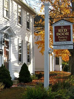 Sign for The Red Door Pottery Studio, located in a white wooden building with trees and autumn leaves in the background.