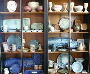 A wooden display cabinet with four shelves filled with various ceramics, plates, bowls, and mugs in pastel and earth tones.
