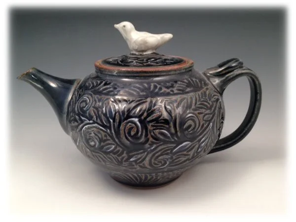 A decorative teapot with a glossy dark finish featuring swirling patterns, topped with a white ceramic bird figurine on the lid handle.