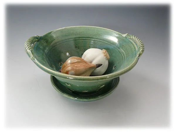 A green ceramic bowl with a matching lid, containing a garlic bulb, a gently peeled onion, and a shallot.