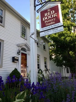Sign for The Red Door Pottery Studio above a garden with purple flowers in front of a white two-story building.