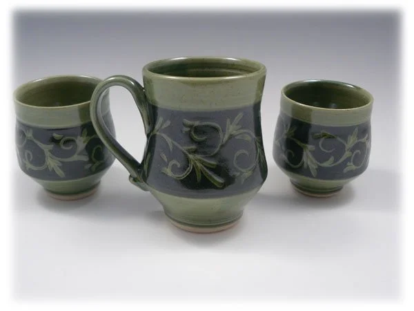 Three ceramic mugs with green and dark blue swirl and vine pattern, one larger and two smaller, placed on a white surface.